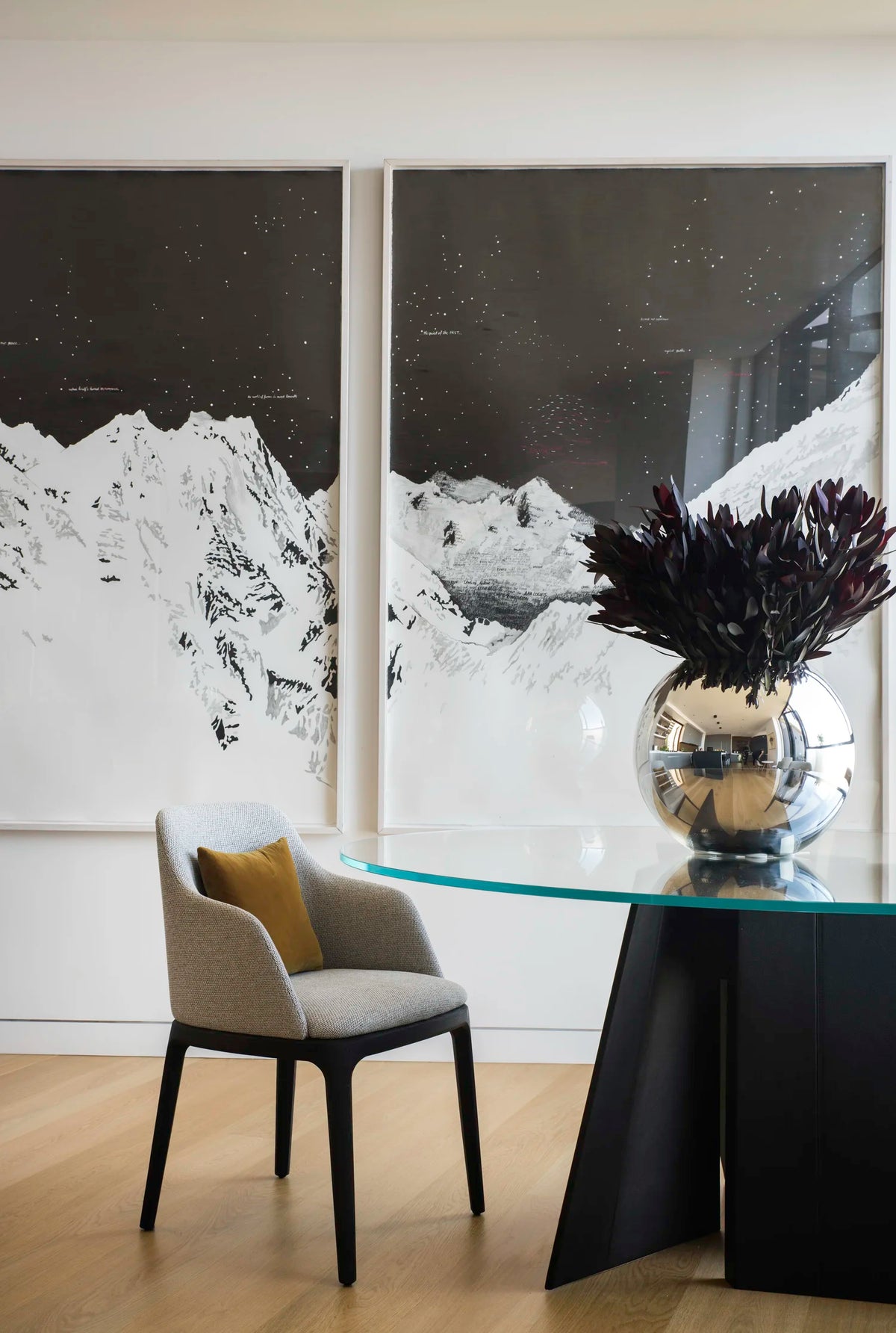 Modern interior with a round glass table and chair, featuring large mountain paintings on the wall.