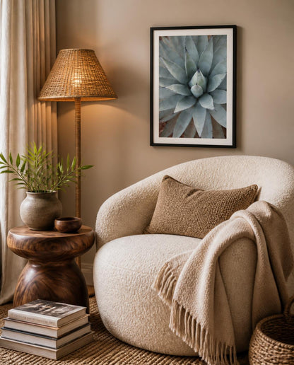 A cozy interior scene with a cream boucle armchair, beige throw blanket, woven floor lamp, potted plant on a wooden side table, and framed Agave botanical fine art photography wall art above the chair. Framed photography by Amanda Anderson. 