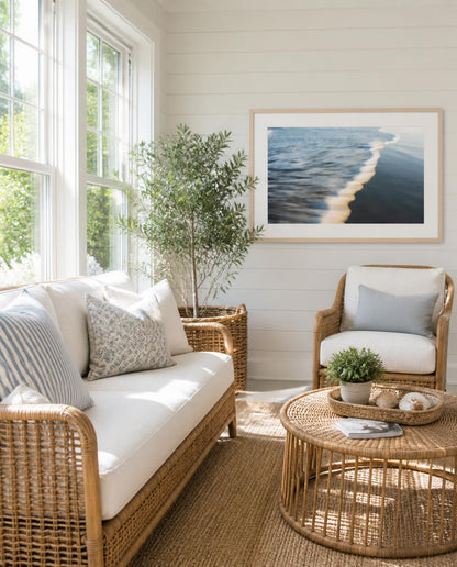 A bright sunny coastal-style living room with a wicker sofa, matching armchair, round wicker coffee table, and a framed ocean wave print by Amanda Anderson on the wall.