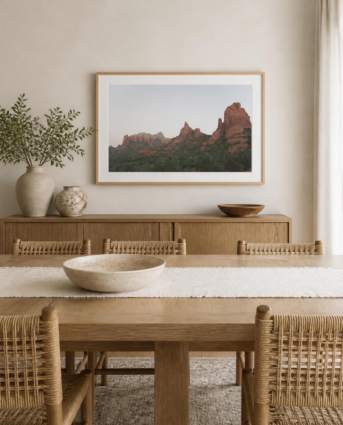 A framed landscape fine art photography print of red rock desert mountains hangs on a beige wall above a wooden sideboard in a styled dining room with woven chairs and a wooden table. Photography by Amanda Anderson. 