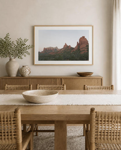 A framed landscape fine art photography print of red rock desert mountains hangs on a beige wall above a wooden sideboard in a styled dining room with woven chairs and a wooden table. Photography by Amanda Anderson. 