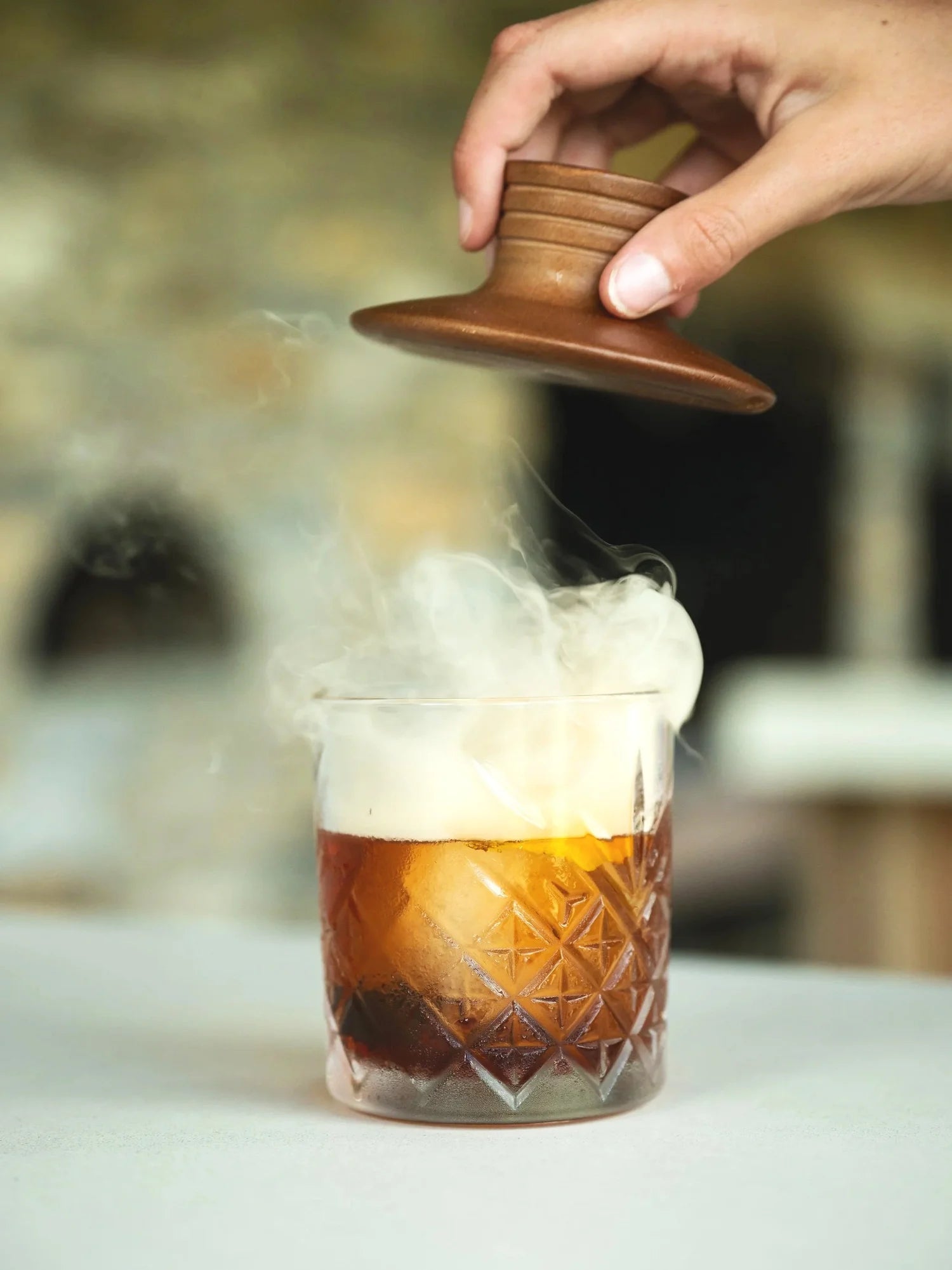 Glass of whiskey with smoke rising from the top and hand holding wooden lid