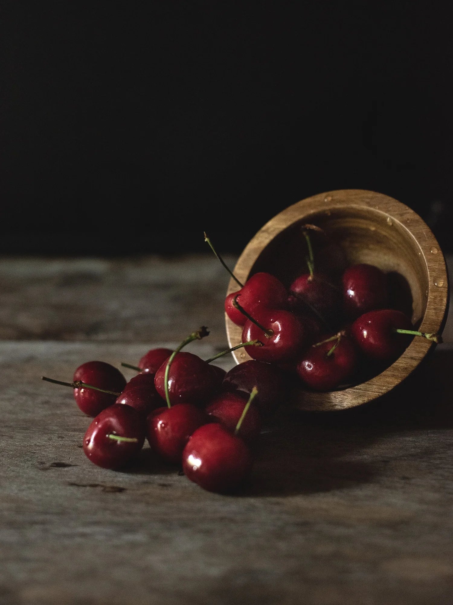 Red cherries spilling out of a wooden bowl on a textured surface with a dark background
