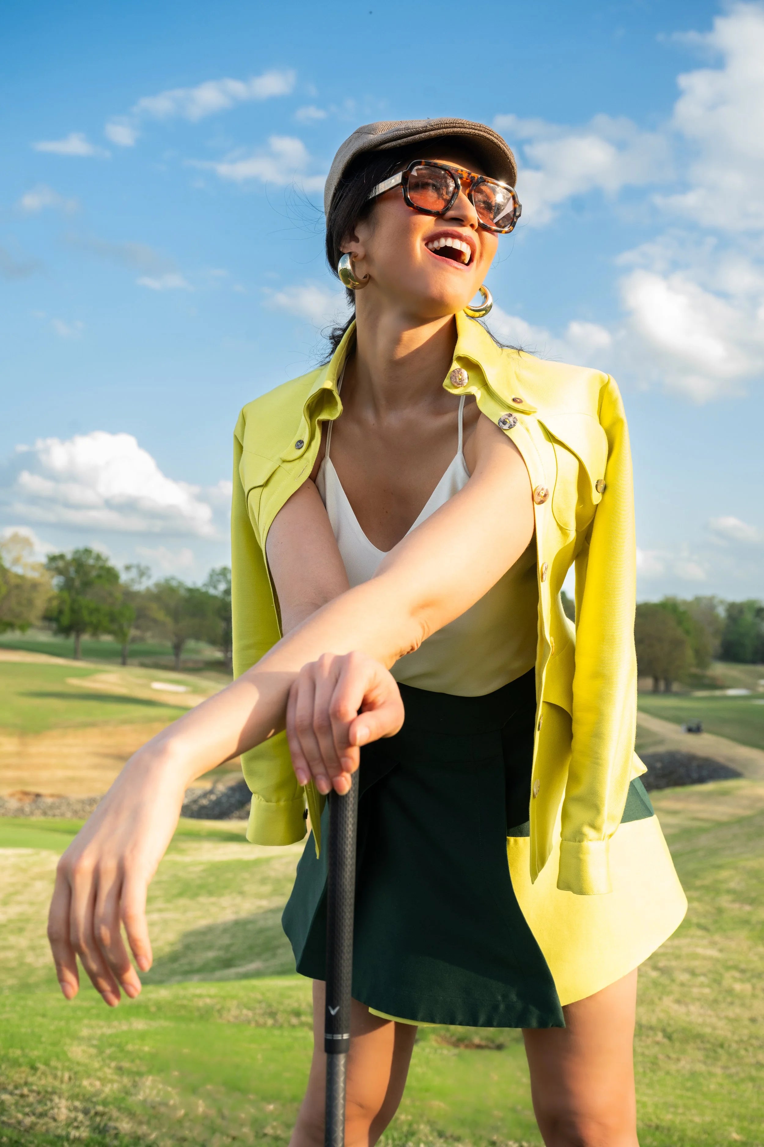 Woman in a yellow jacket and green skirt holding a golf club on a golf course.