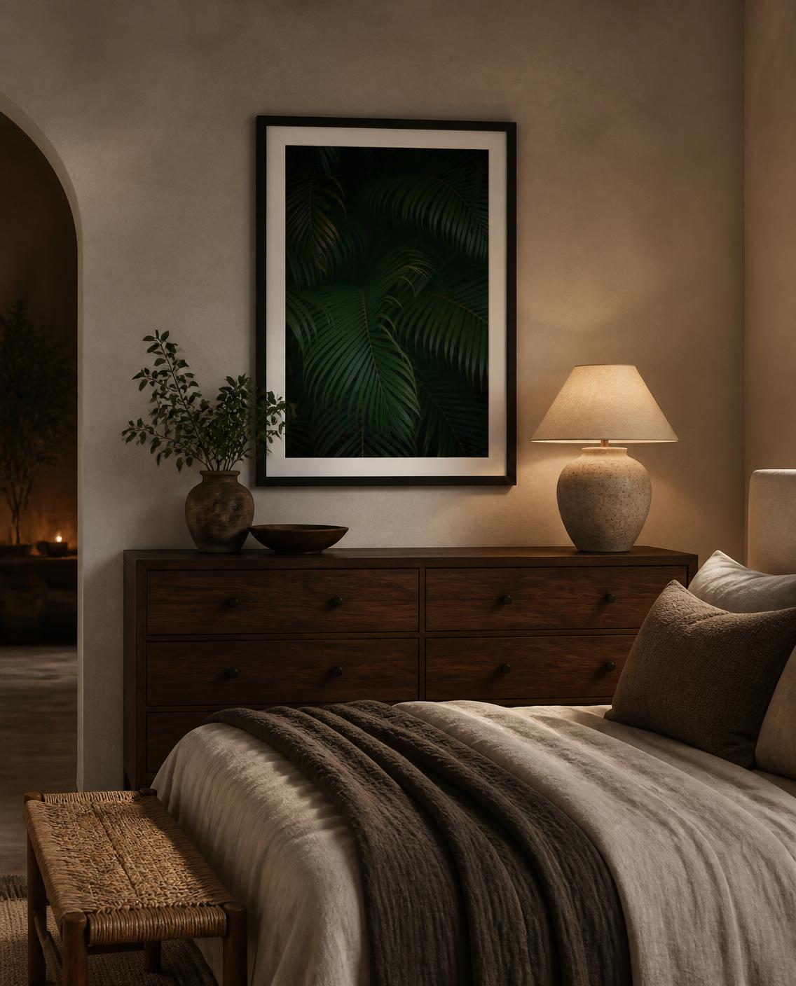 A framed dark green tropical palm leaf fine art photography print hangs on a textured beige wall above a wooden dresser in a cozy, dimly lit bedroom with Arhaus vibes. Photo taken by Amanda Anderson.