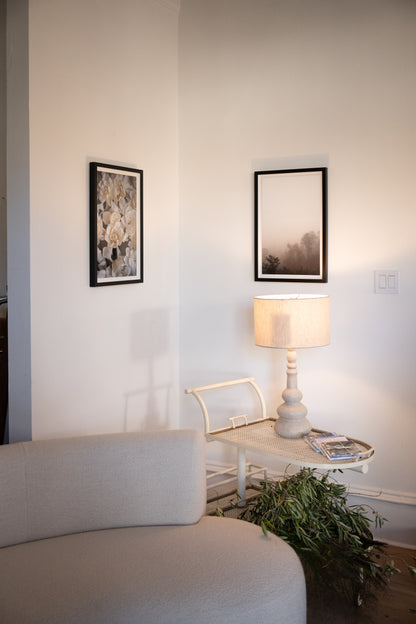 A styled interior corner featuring a cream upholstered sofa in the foreground, a small white side table with a lit table lamp, framed wall art, and a green plant on the floor against a warm off-white wall. Photography prints by Amanda Anderson