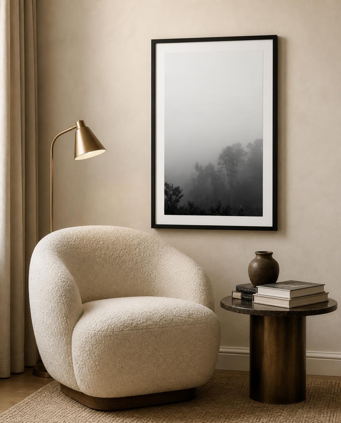A framed black and white landscape photograph of a foggy forest hangs on a beige wall in a styled room with a cream boucle armchair, a brass floor lamp, and a small side table. Photography by Amanda Anderson. 
