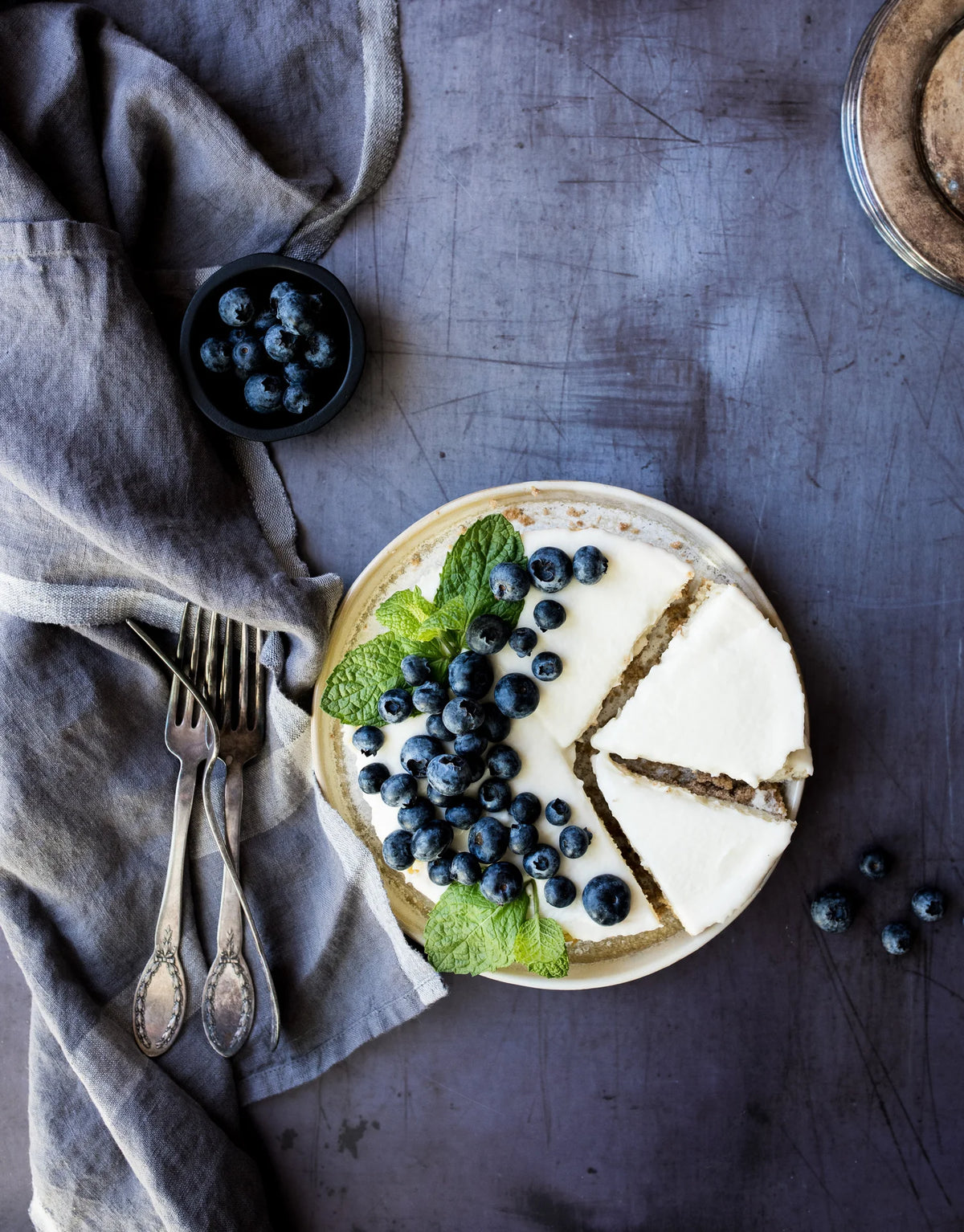 Blueberry cake with a slice taken out on a dark surface with a gray cloth and utensils.
