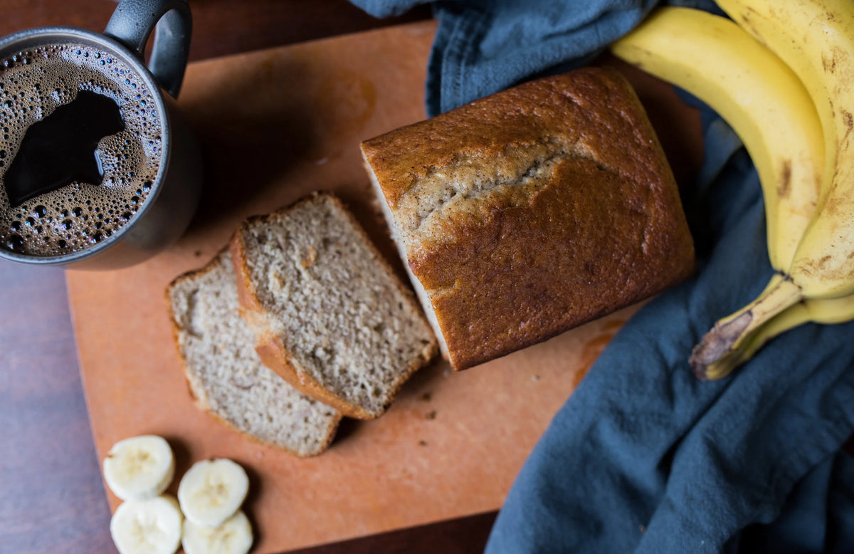 Loaf of banana bread with slices, a cup of coffee, and bananas on a wooden board.
