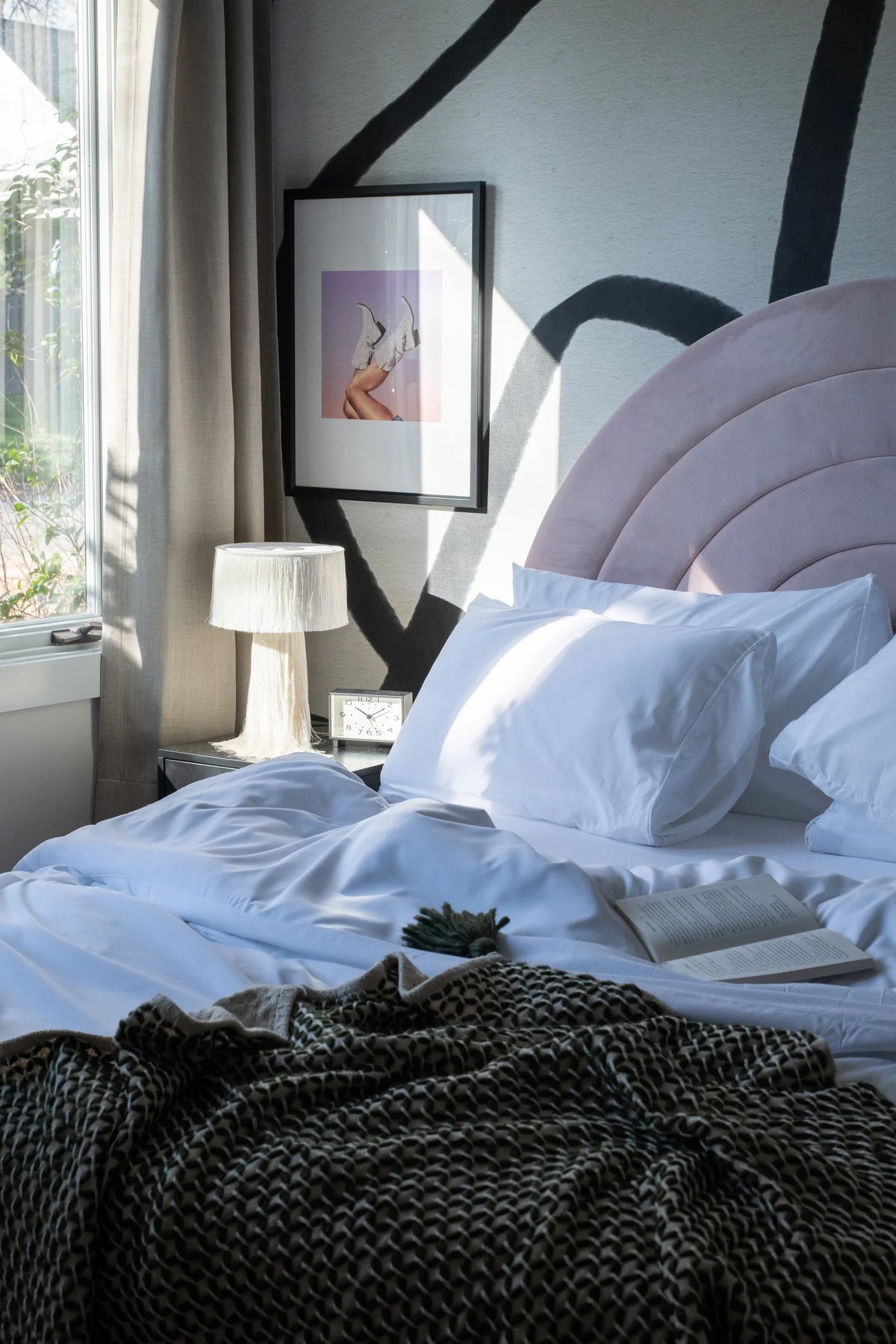 Bed with white bedding and a tufted blush headboard with a nightstand, lamp, clock, and book
