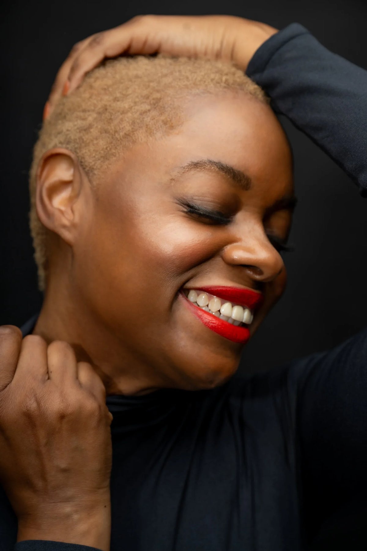 Woman with short hair and red lipstick smiling with hands around her head against a dark background