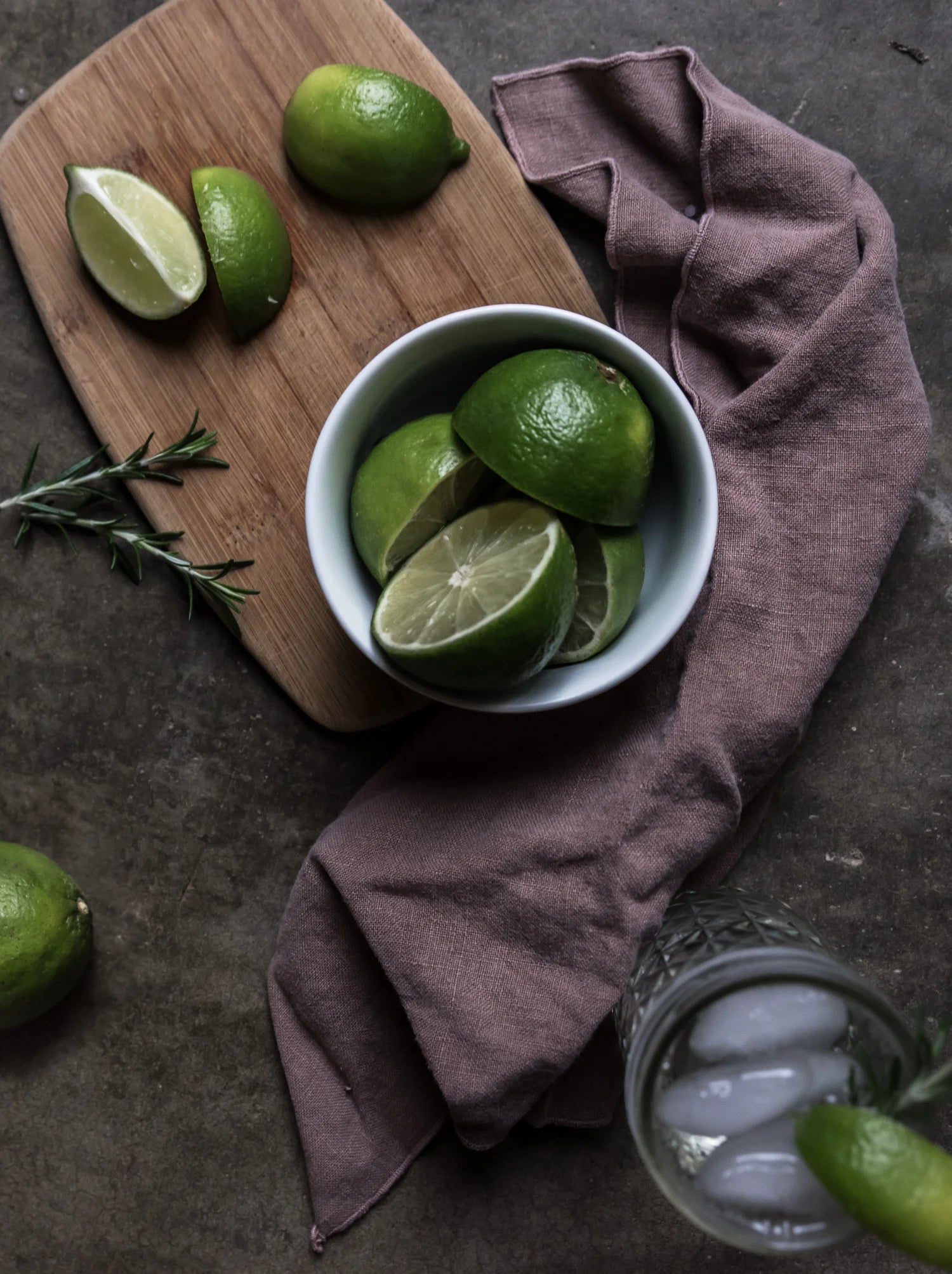 Limes in a bowl on a wooden cutting board with a purple cloth and glass of ice in the background.