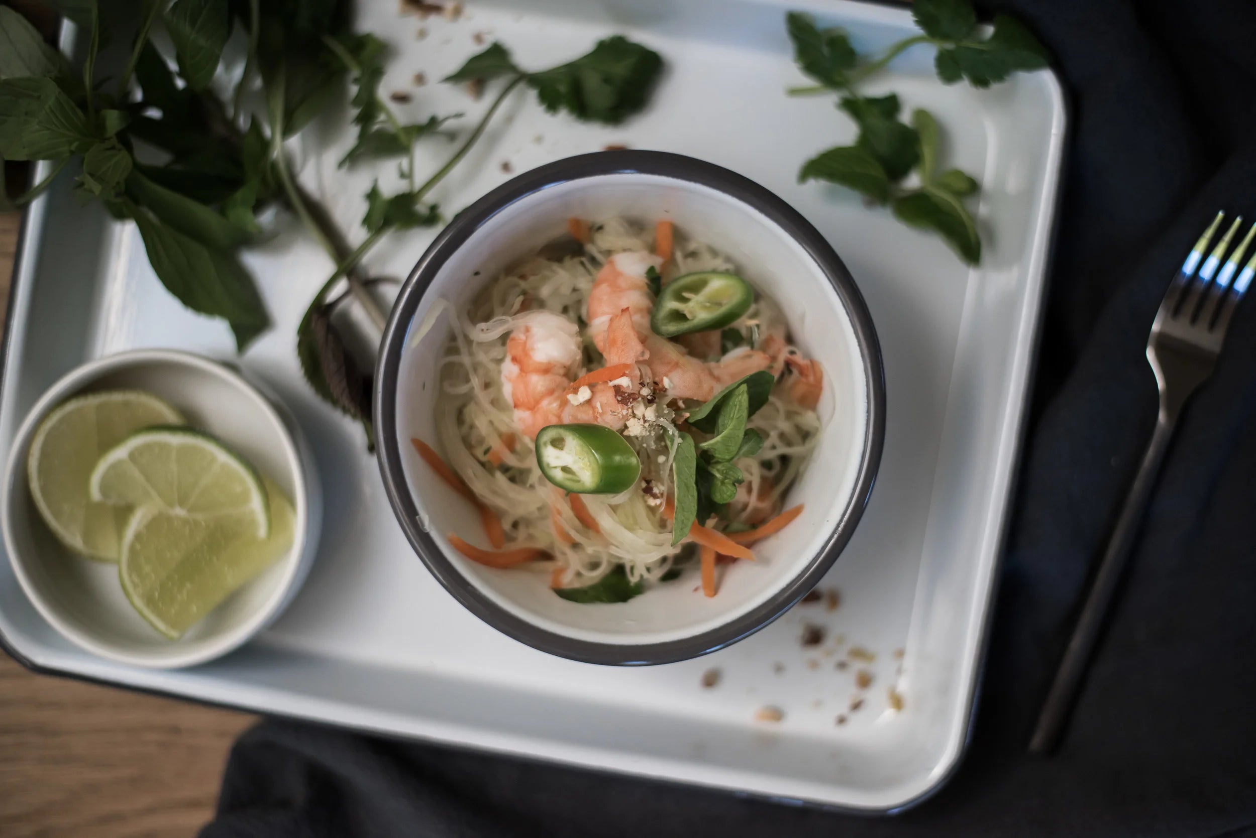 Bowl of ramen with shrimp and vegetables on a white tray with lime wedges and a fork.