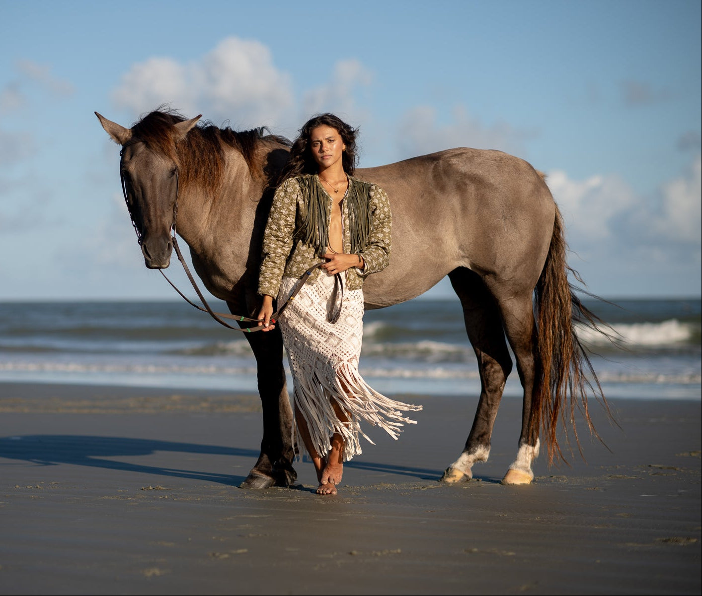 Fashion photographer, Amanda Anderson, photographed a model, Sydney Schertz, on a beach next to a large brown horse, wearing a patterned green jacket designed by McNeely Purcell with a white fringed skirt. The ocean and blue sky are visible in the background.
