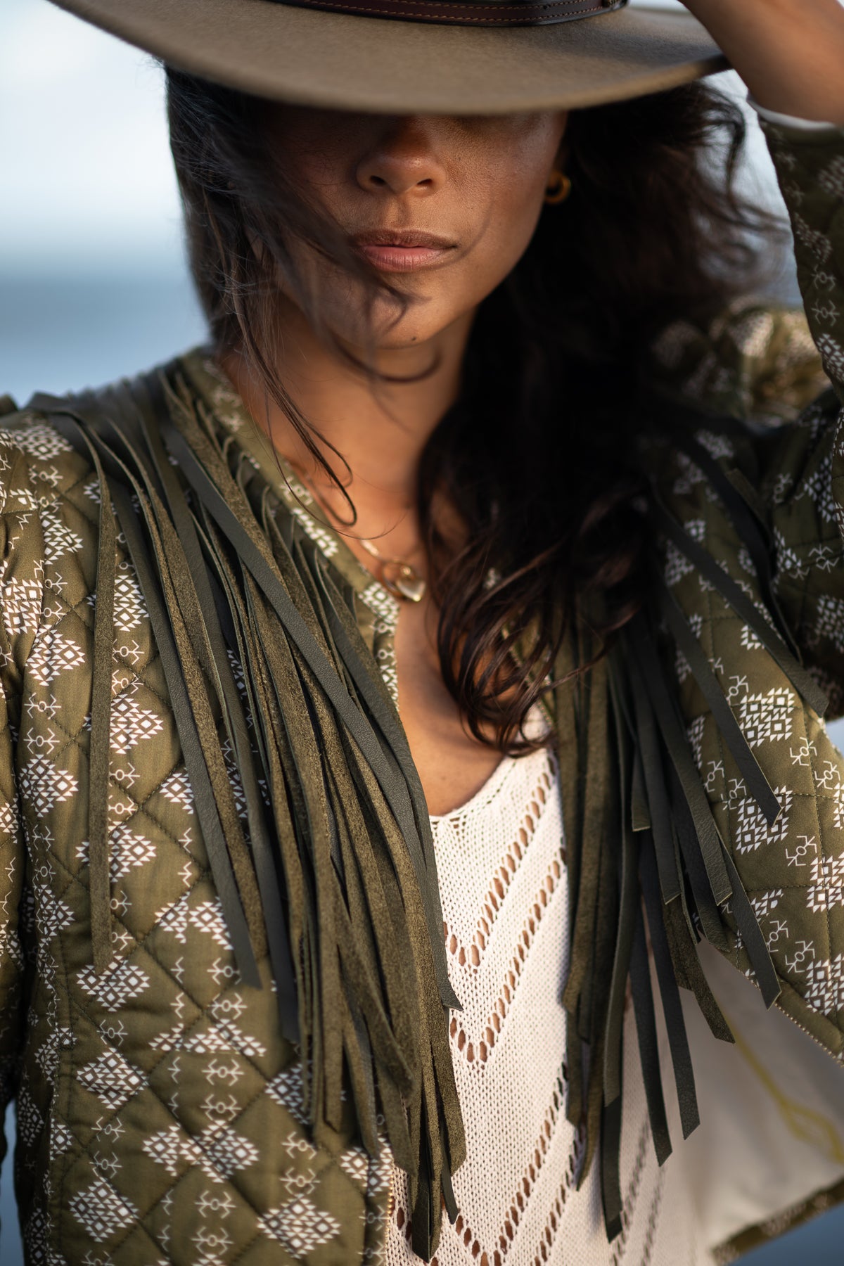A close-up lifestyle shot of a model wearing a wide-brimmed brown hat, a white knit top, and an olive green fringed jacket with a geometric pattern designed by McNeely Purcell was photographed by commercial photographer, Amanda Anderson on a beach in South Carolina.