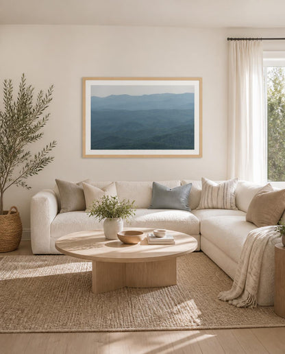 A framed landscape fine art photography print depicting blue mountain layers, hung on a beige wall above a modern white sectional sofa in a bright, neutral-toned living room. Photography by Amanda Anderson.
