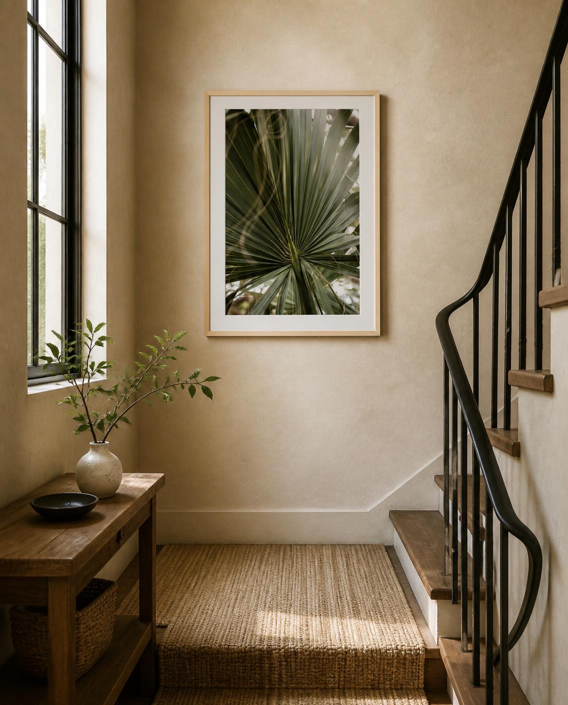 A framed botanical art print of a green palm leaf hangs on a beige wall in a styled staircase hallway with a wooden console table and a woven rug. Photo by Amanda Anderson.