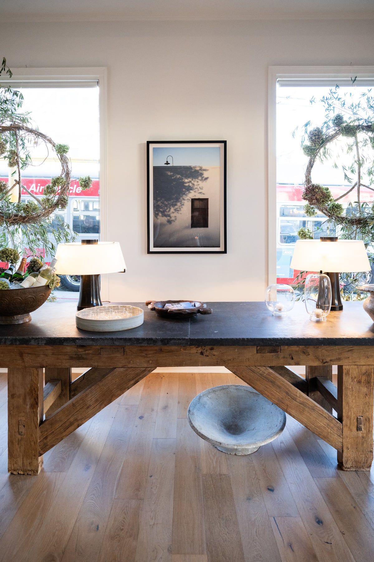 A rustic wooden dining table with a dark stone top is styled in a bright room with two windows, framed wall art, table lamps, and decorative wreaths showcases a framed fine art photo on the wall by Amanda Anderson.