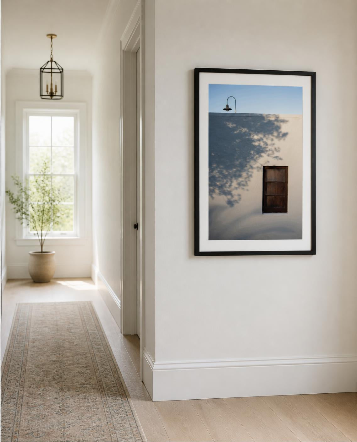 A framed fine art photography print hangs on a white wall in a bright, modern hallway. The print features a minimalist photograph of a blue sky, a white wall, and a dark window, enclosed in a black frame with a white mat. Photo by Amanda Anderson
