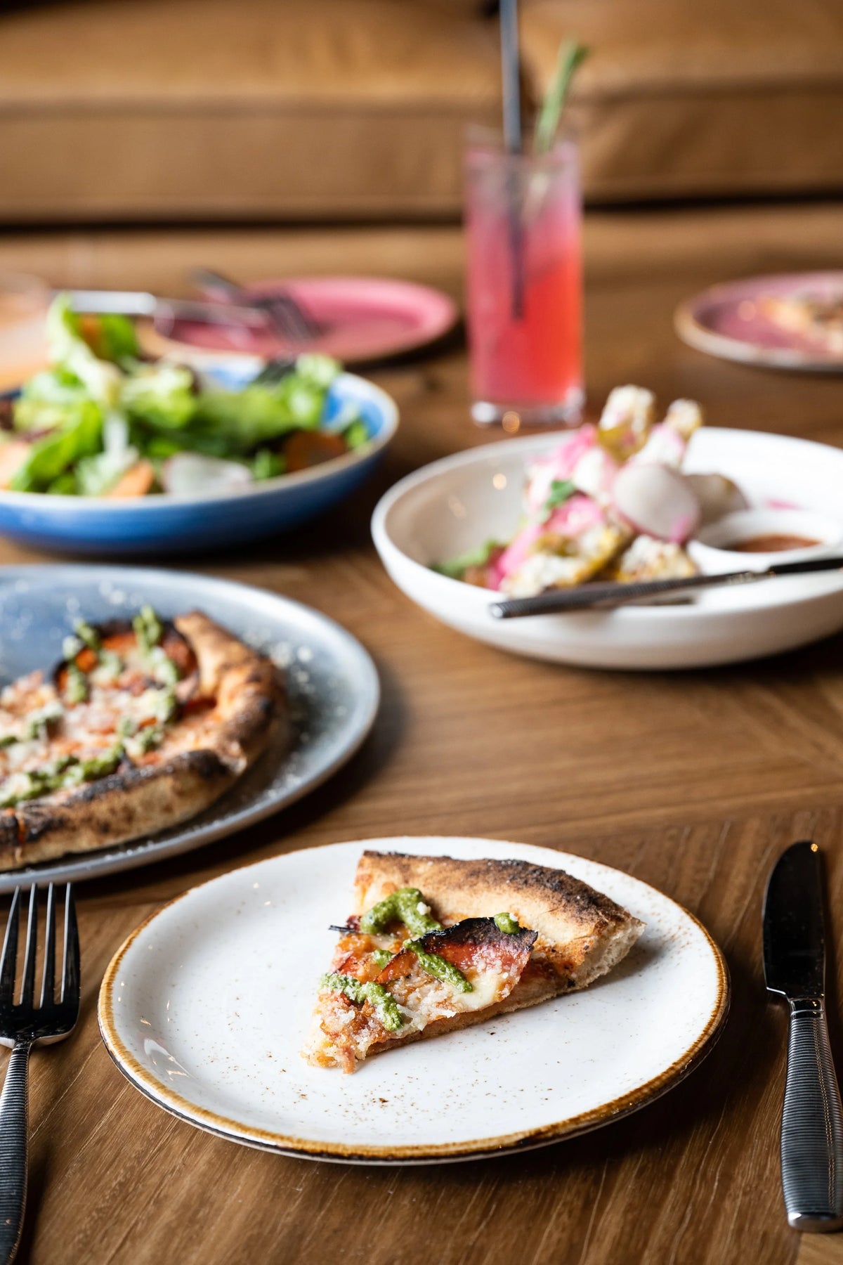 Slice of pizza on a plate with a salad and drinks in the background on a wooden table.