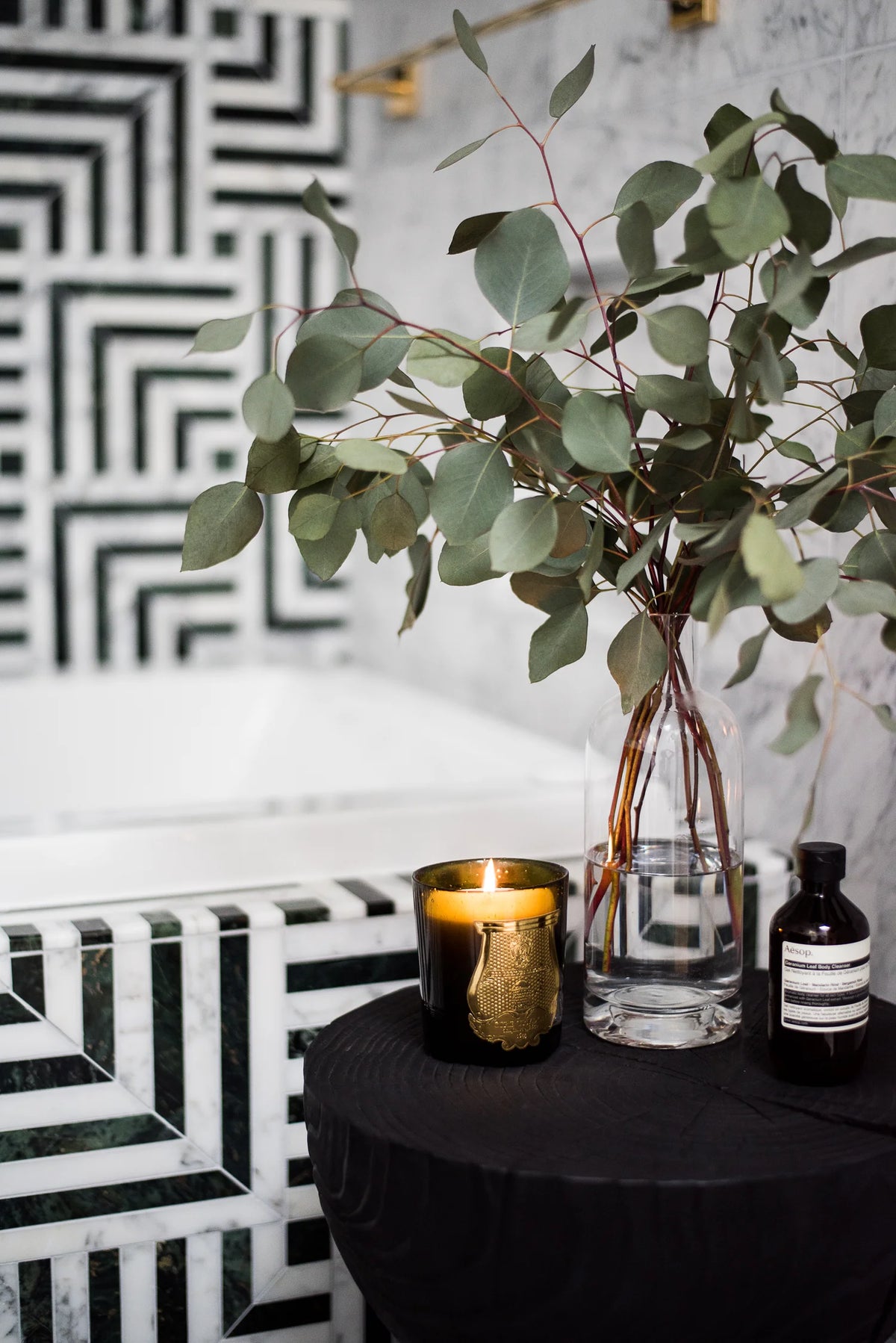 Decorative setting with a vase of greenery and candle next to a black and white geometric patterned bathtub.