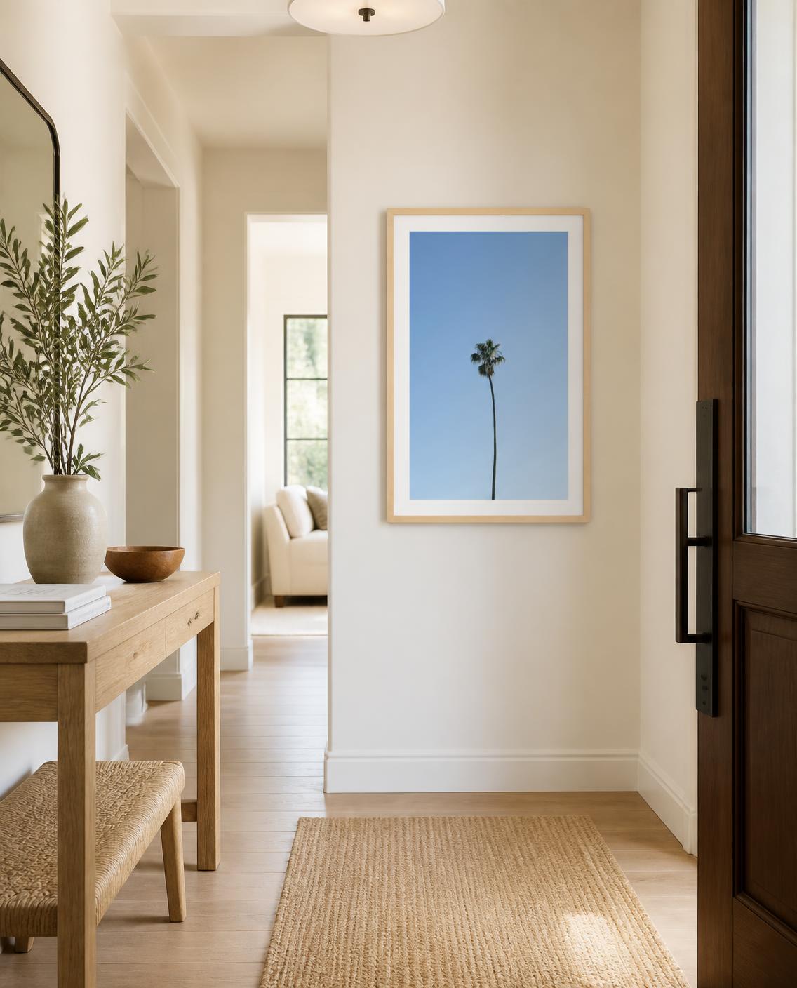 A framed art print featuring a minimalist blue sky with a single palm tree hangs on a beige wall in a bright, modern hallway with light wood floors and a woven rug. Photography by Amanda Anderson. 