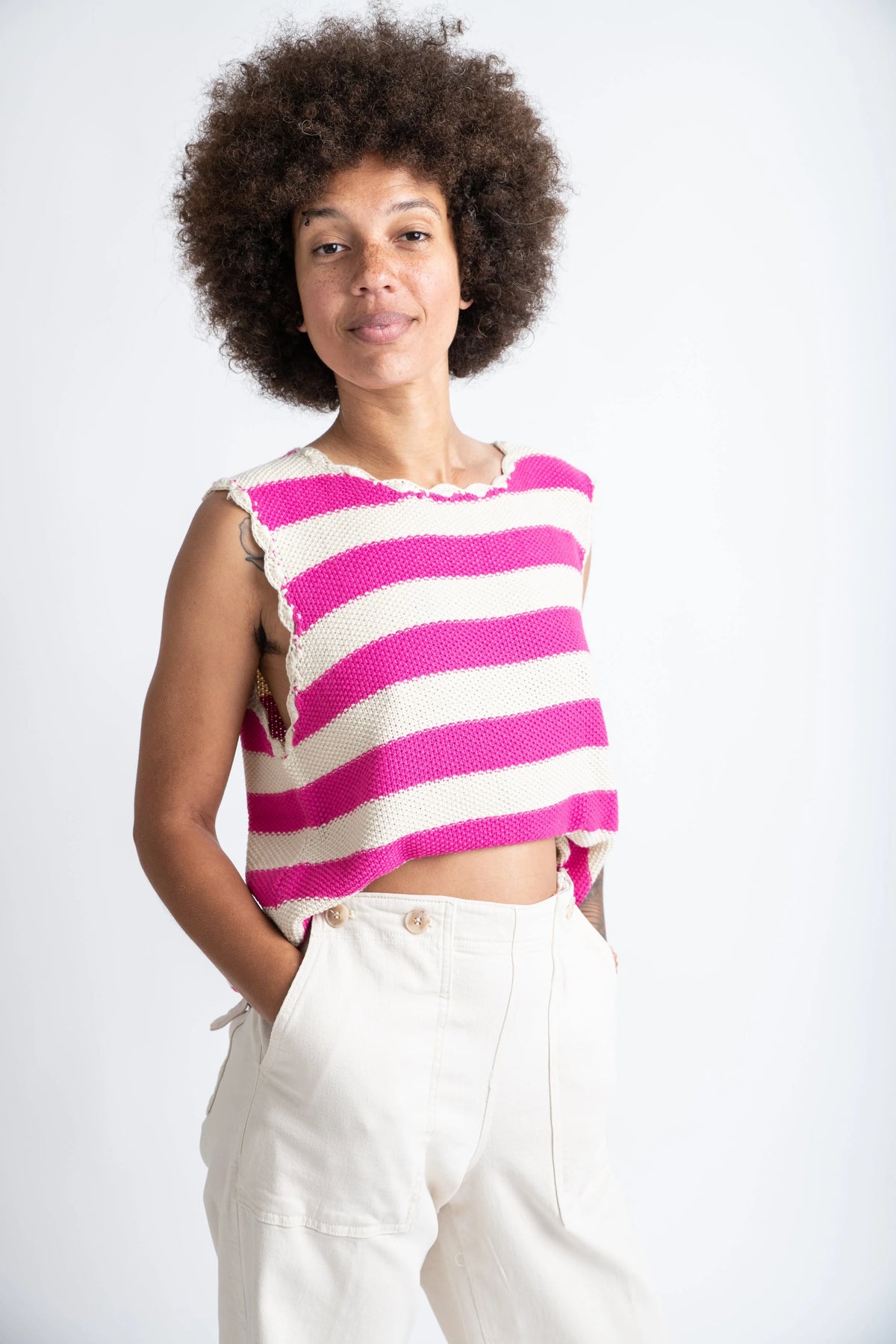 Woman wearing a pink and white striped sleeveless top with white pants on a plain background