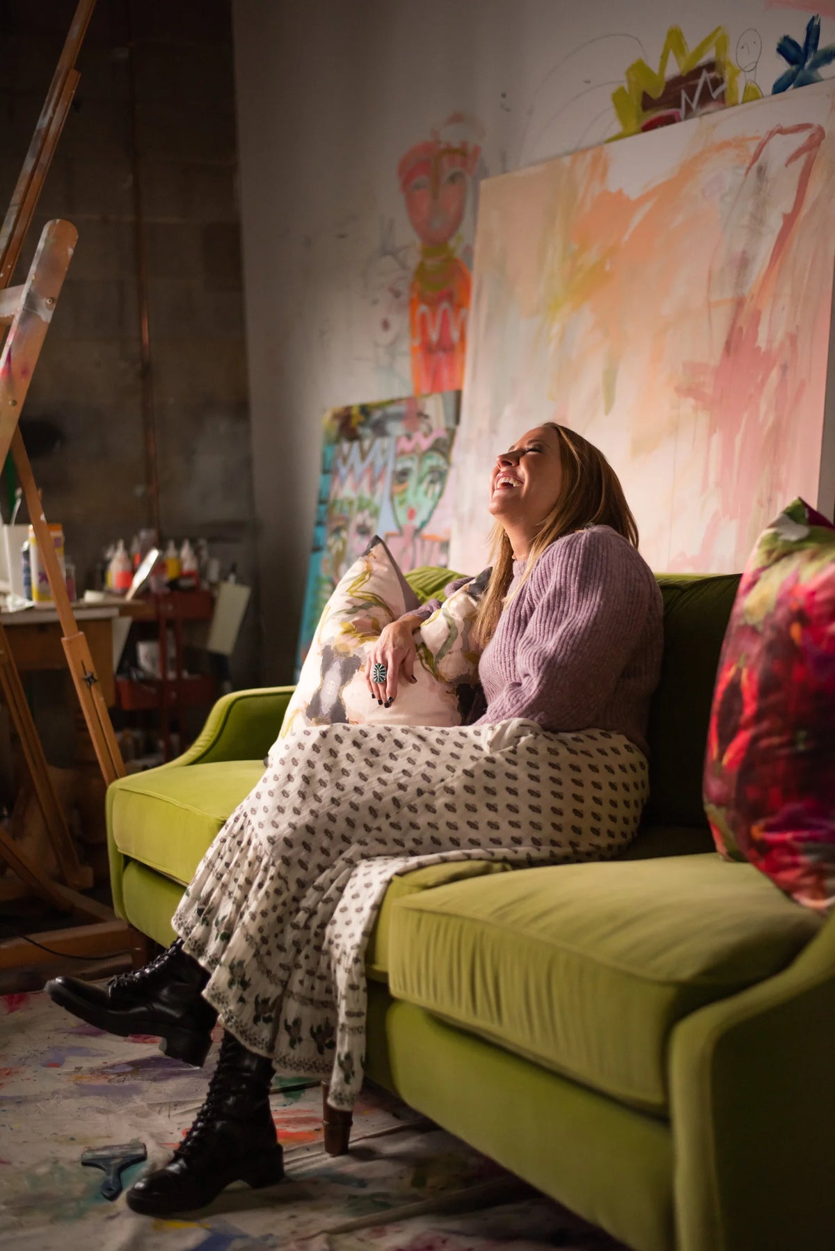 Woman laughing while sitting on a green couch in a room with artwork and a painting easel