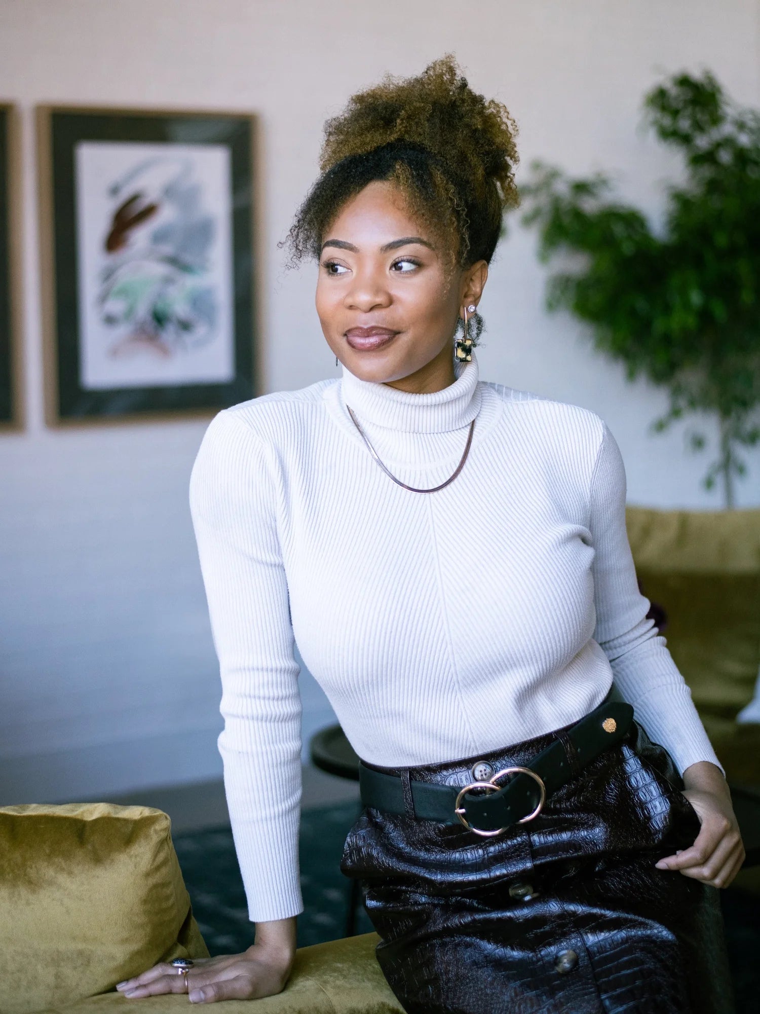 Woman wearing a white turtleneck and black skirt sitting in a room with a plant and framed picture in the background.